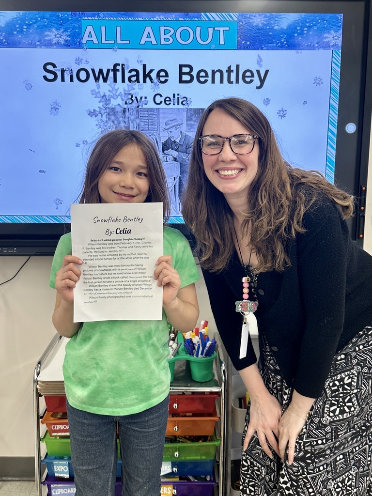 A student stands beside a teacher holding a report about Snowflake Bentley, smiling in front of a classroom display.