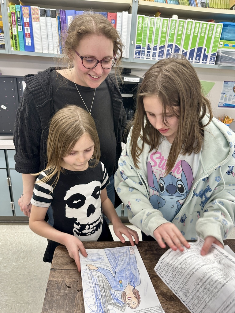 A teacher leans over two students reviewing a drawing and worksheet together at a desk in a classroom.