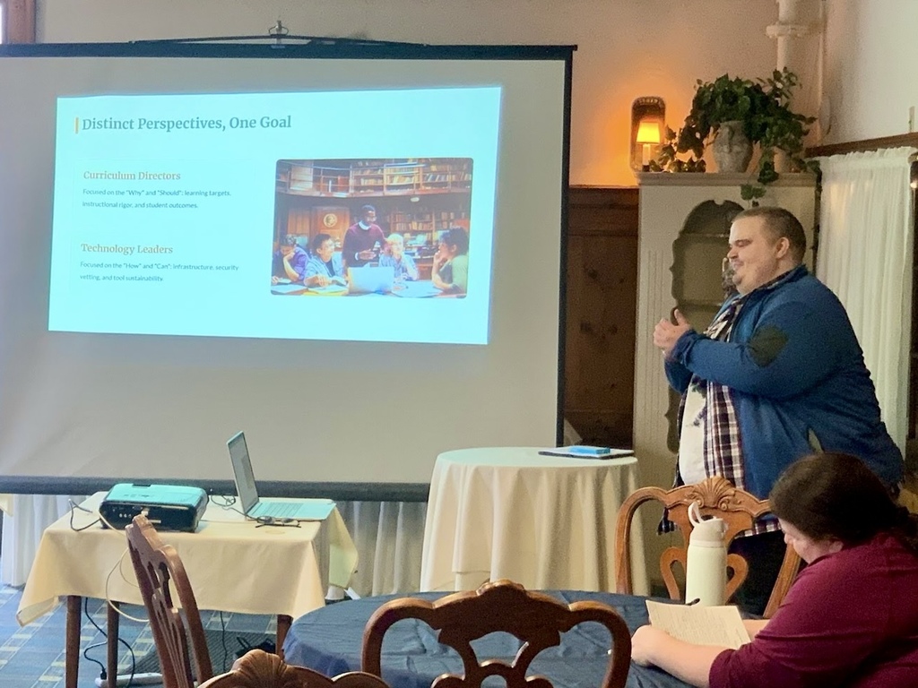 A man stands at the front of a small meeting room presenting beside a projection screen displaying a slide titled “Distinct Perspectives, One Goal.” A laptop and projector sit on a table, while attendees are seated at round tables listening.