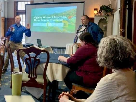 Two presenters stand at the front of a meeting room leading a session titled “Aligning Mission & Expertise: The AI Frontier in Vermont,” projected on a screen. Several attendees sit at tables facing them, listening and taking notes.