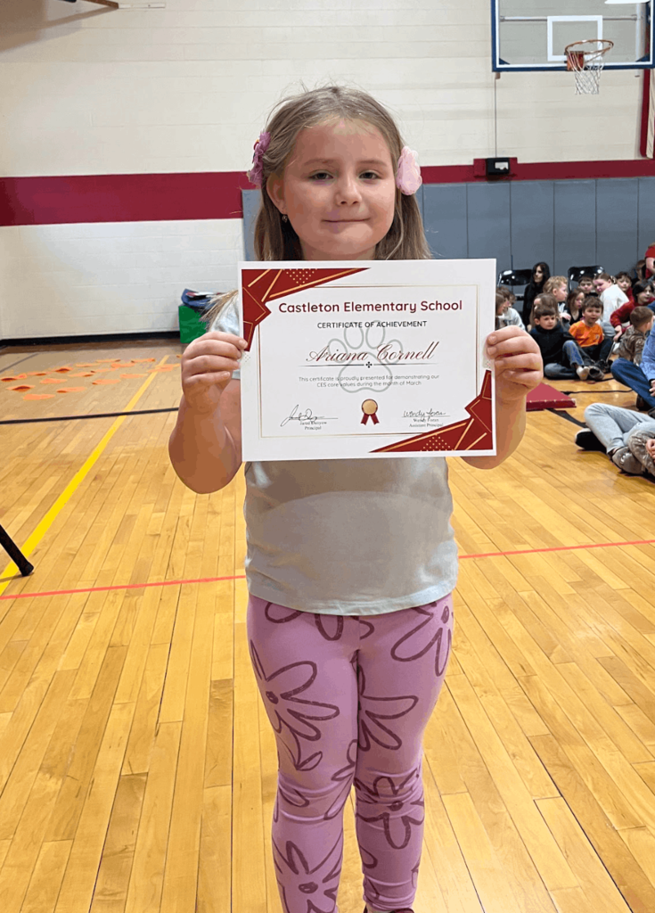 Student holds a “Castleton Elementary School Certificate of Achievement” while standing in a gym with classmates seated in the background.