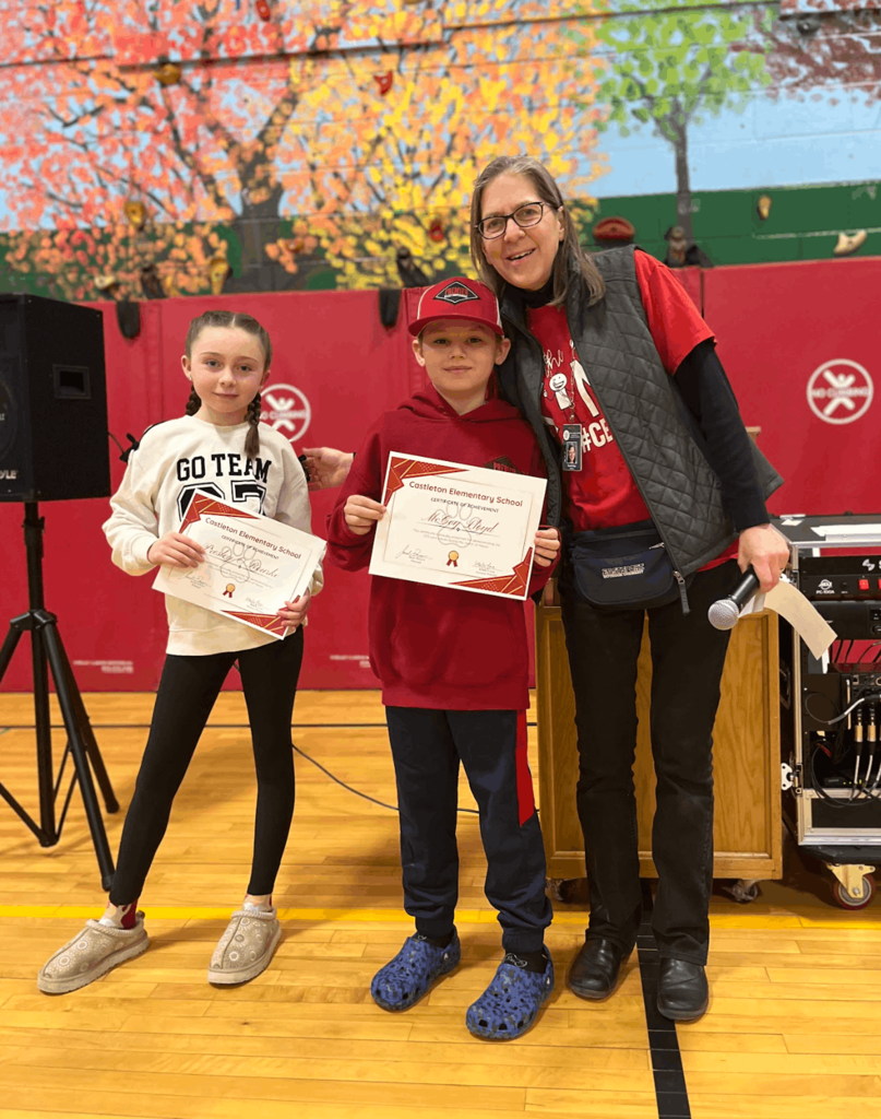 Staff member stands with two students holding certificates in a gymnasium during an awards ceremony.