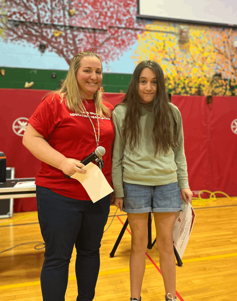 Staff member stands with a student holding a certificate while speaking into a microphone in a gym.