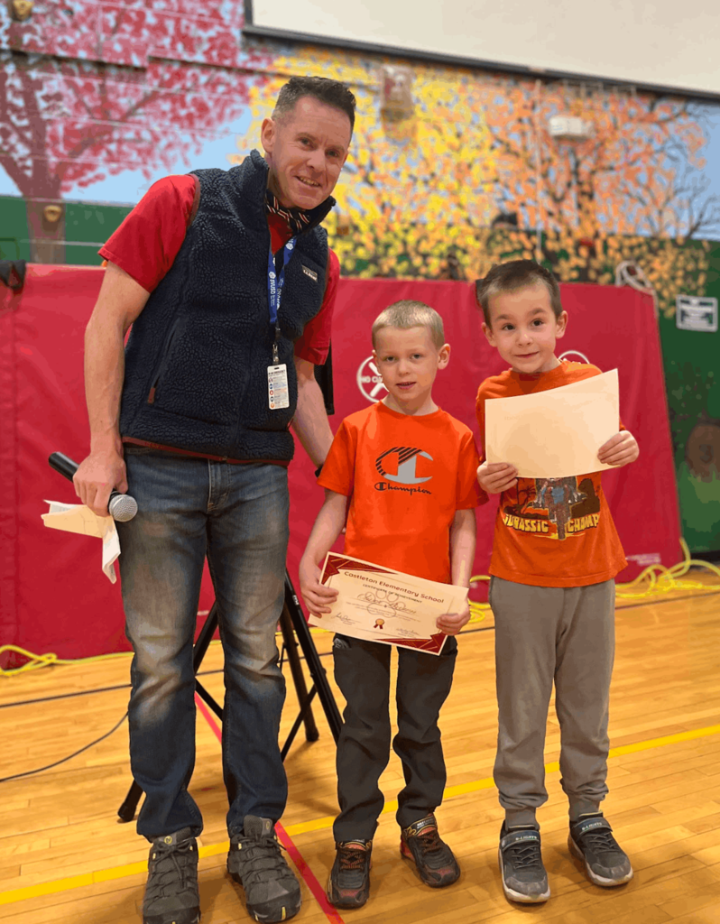 Adult with microphone stands with two young students holding certificates in a school gym during an awards presentation.