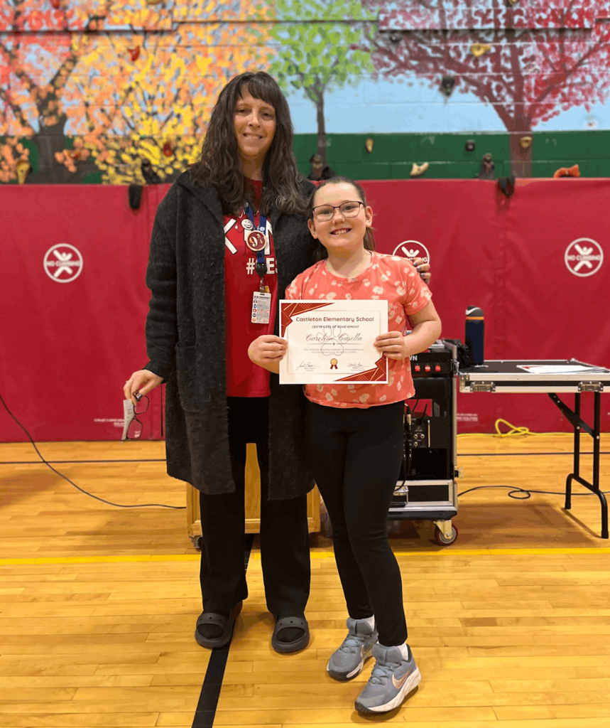 Staff member stands with a student holding a certificate, smiling during a school assembly.