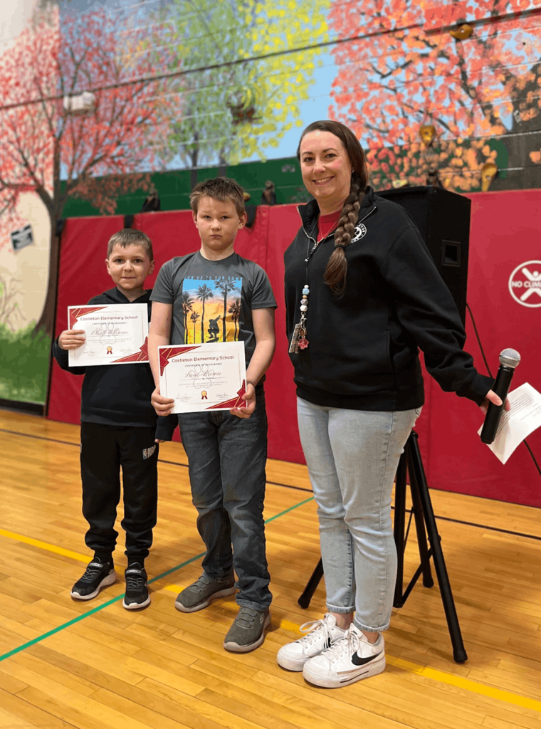 Staff member stands with two students holding certificates in a gym, posing in front of a colorful mural.