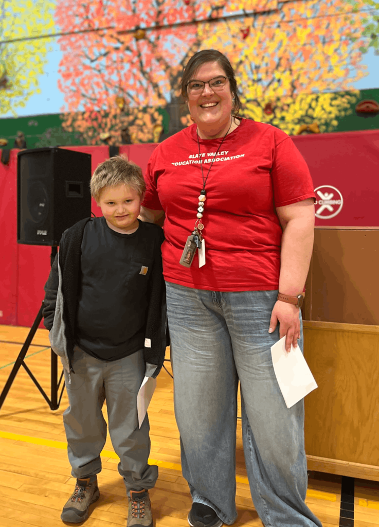 Staff member in a red shirt stands beside a student holding a certificate during a school recognition event.