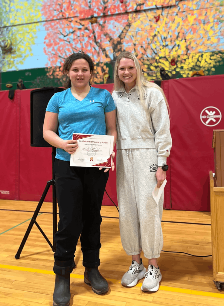 Staff member stands with a student holding a certificate, posing in front of gym equipment and a mural.