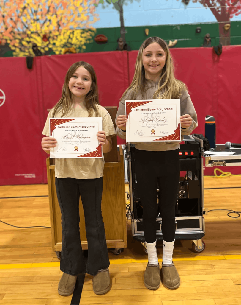 Two students stand side by side holding certificates during a school awards ceremony.
