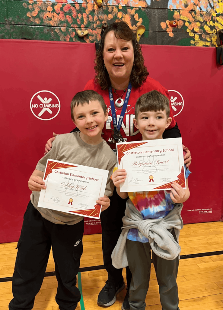 Staff member stands behind two students holding certificates, smiling during a school awards presentation.