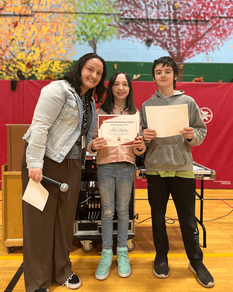 Staff member stands with two students holding certificates during a recognition event in a gym.