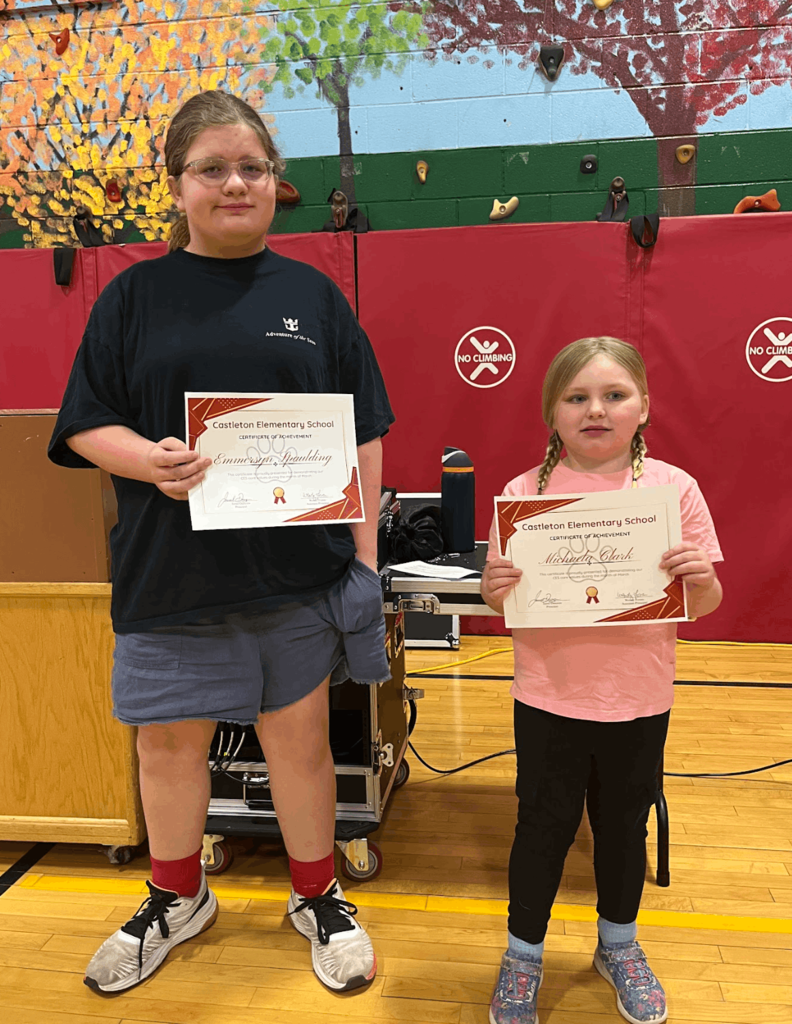 Two students stand holding certificates in a gym during a recognition event.