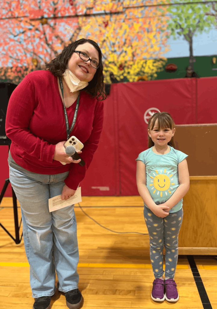 Staff member stands with a student holding a certificate in a gym during a school recognition event.