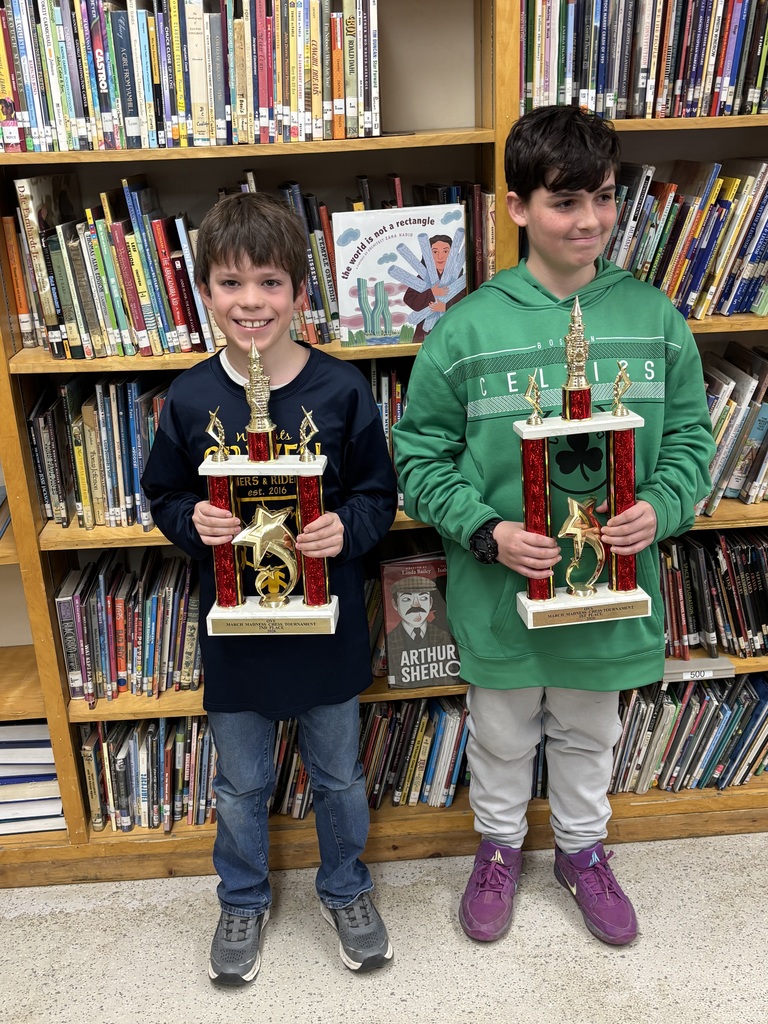 Two students stand in a school library holding large red-and-gold trophies, smiling in front of bookshelves filled with books.
