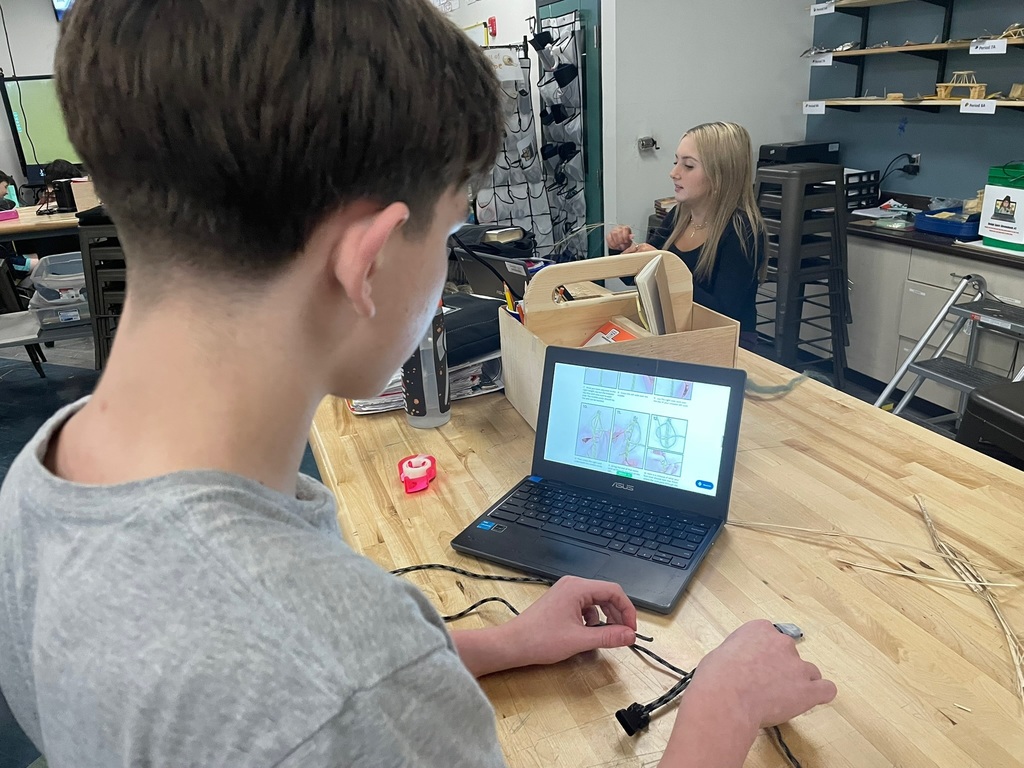 Student following instructions on a laptop while assembling fiber materials for a weaving or cordage project.