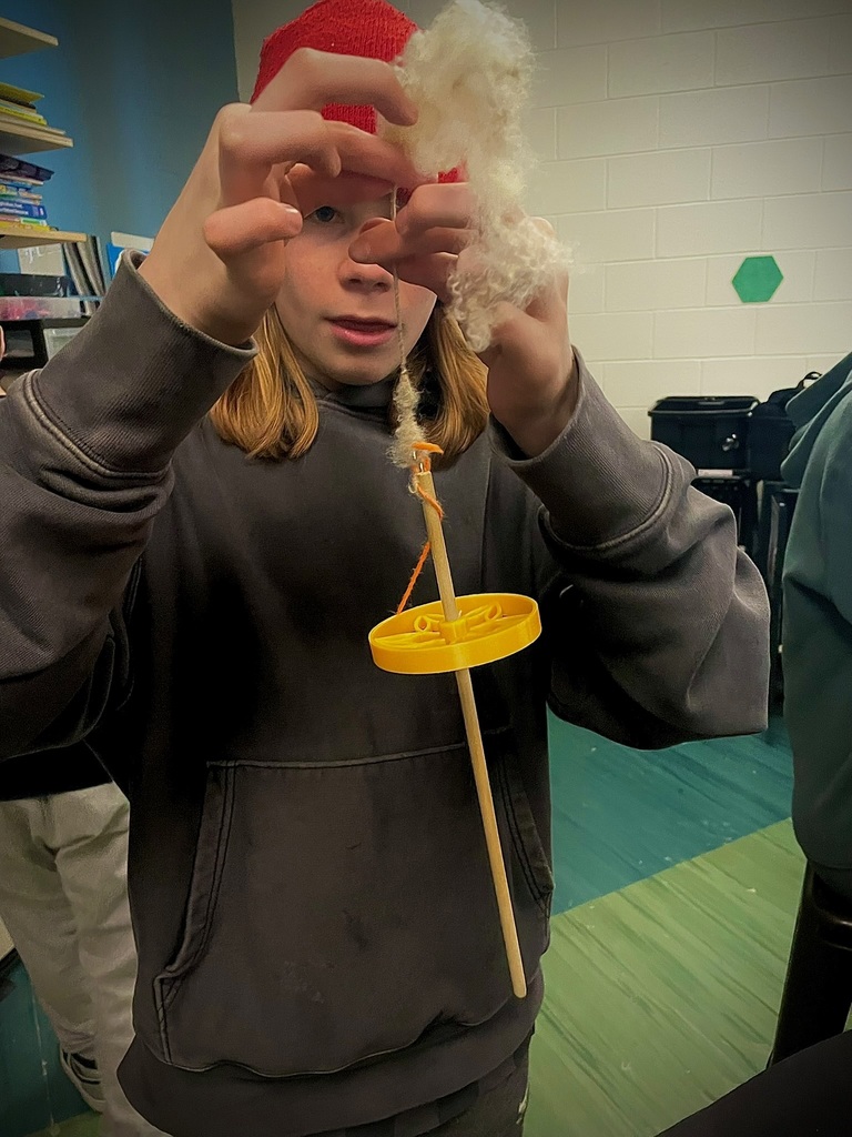 Student using a drop spindle to spin white wool into yarn during a hands-on fiber arts activity.