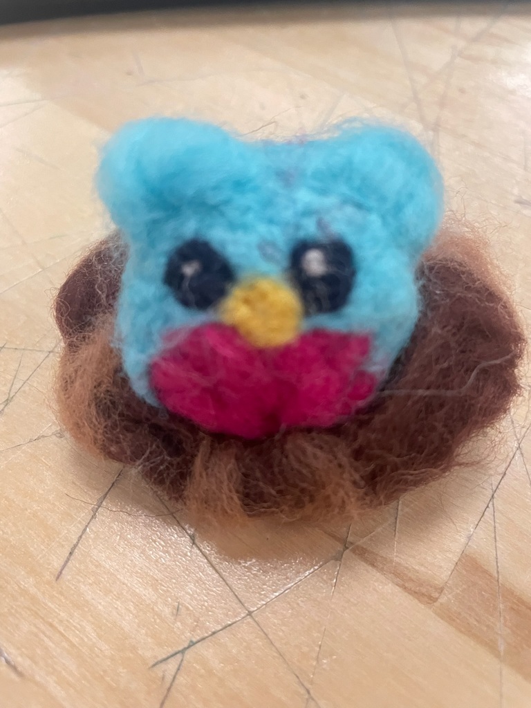 Close-up of a small, colorful needle-felted owl with blue, pink, and brown wool details.