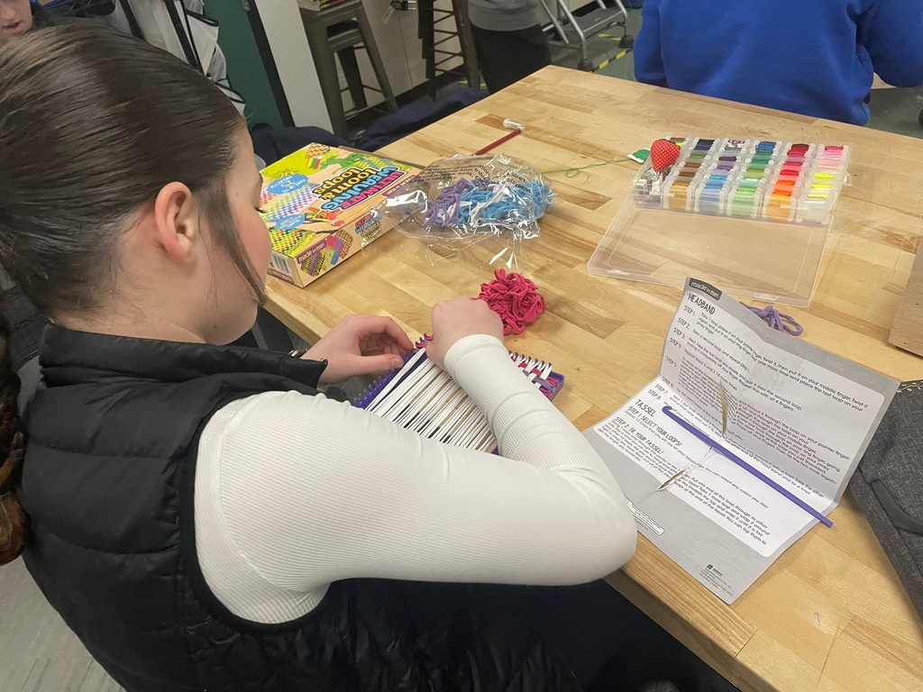 Student using a small loom to weave colorful loops into a textile project on a classroom workbench.