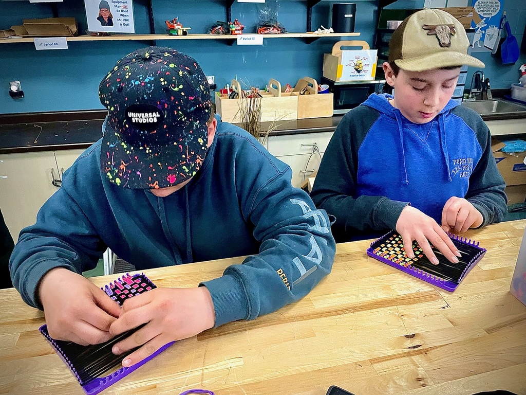 Two students seated at a table weaving on small looms, focusing on their textile projects.