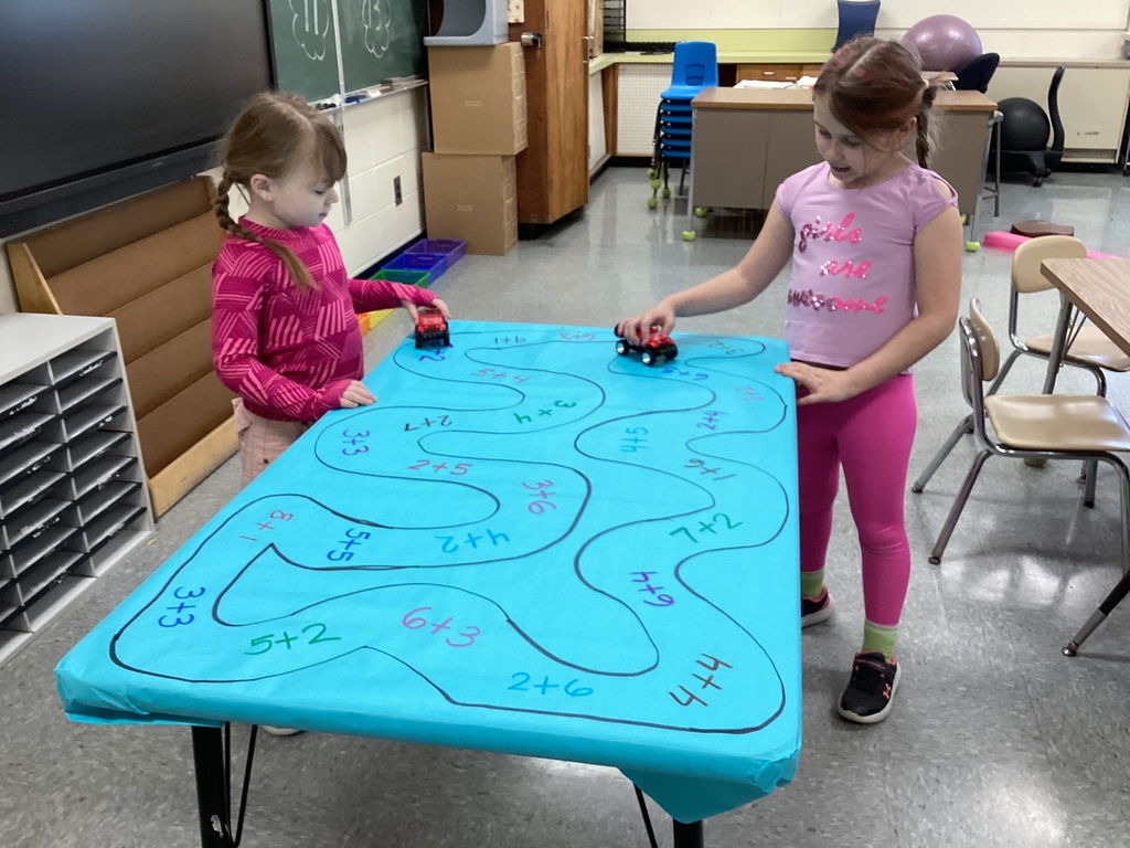 Two students stand at a table moving toy cars along a hand-drawn path of math problems, combining movement with addition practice.