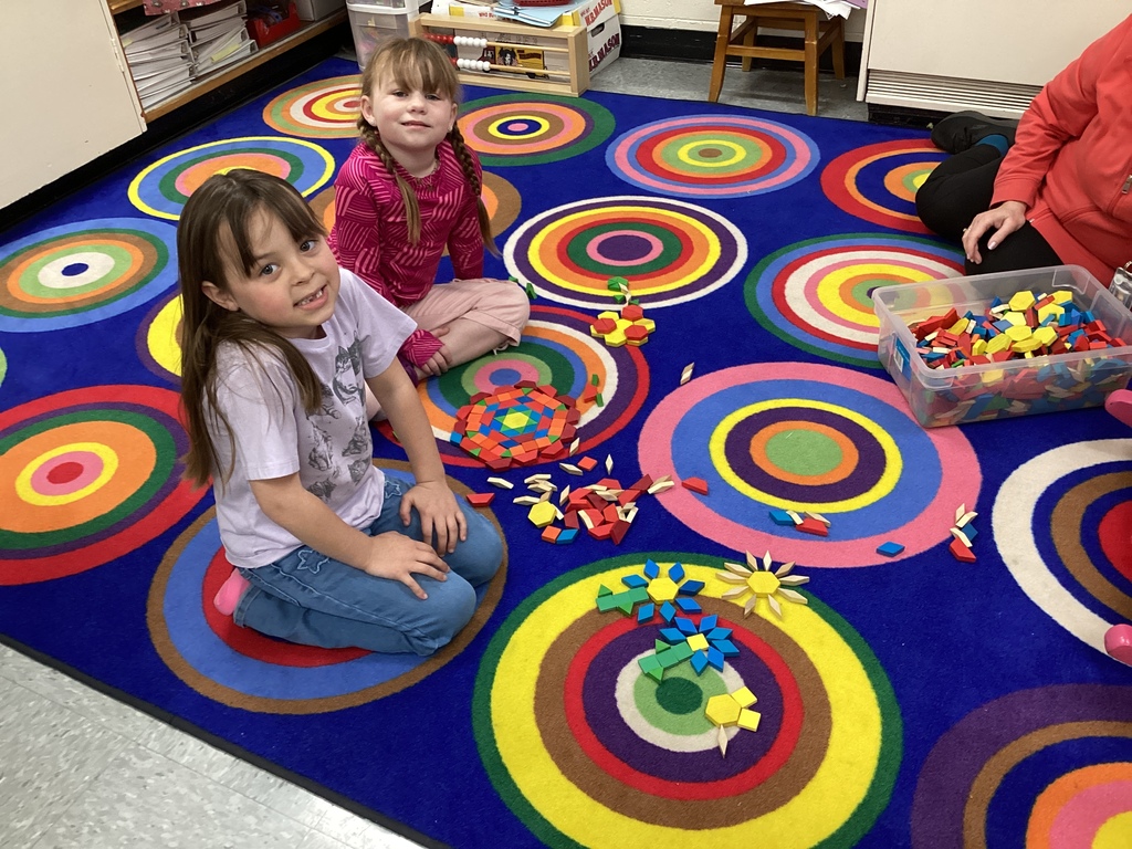 Two first grade students sit on a colorful classroom rug, creating geometric patterns with small wooden blocks spread around them.