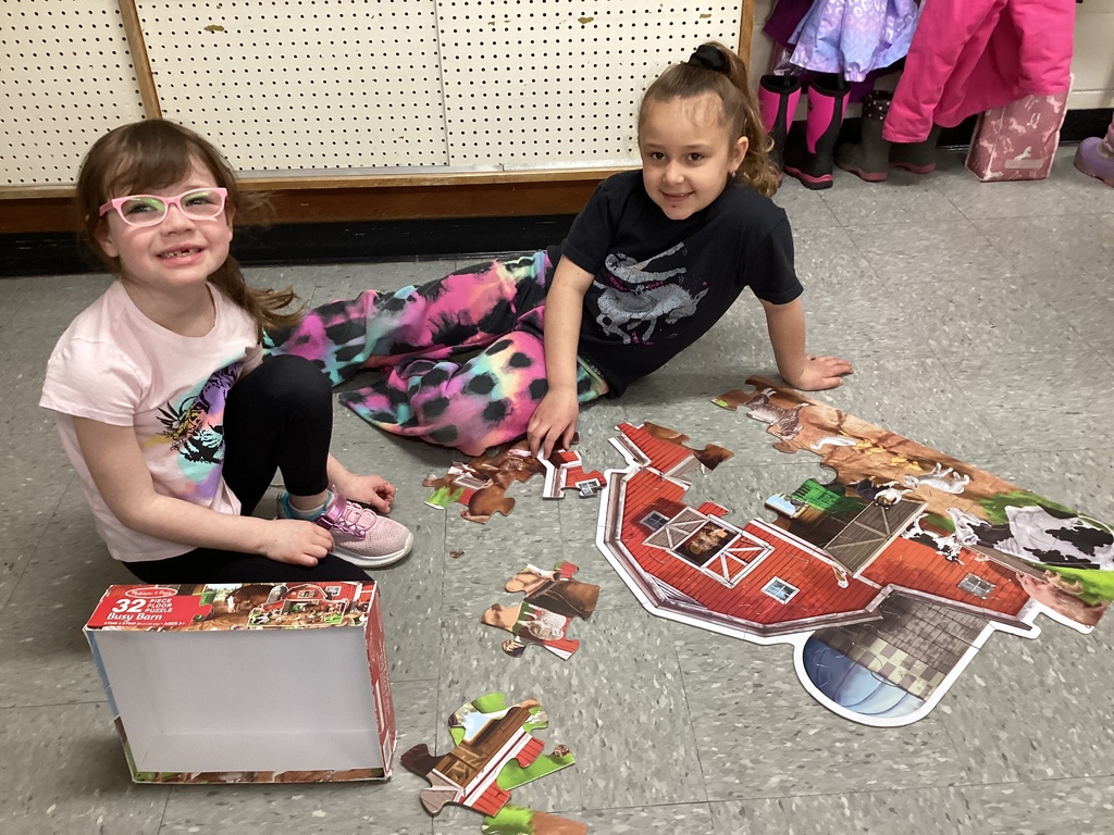 Two students sit on the floor assembling a farm-themed floor puzzle, with puzzle pieces scattered nearby.