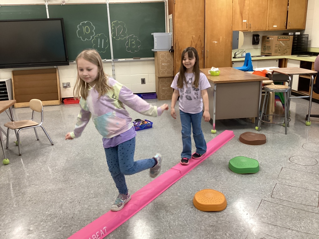 Two students balance and move along a foam beam while navigating a classroom obstacle course.