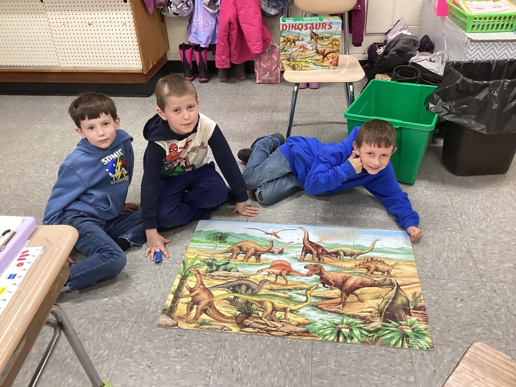 Three students sit around a large dinosaur puzzle on the floor, working together to complete the scene.
