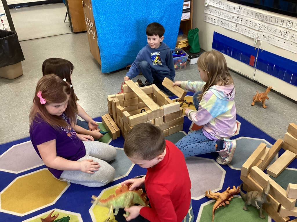 Four students sit on a rug building structures with wooden blocks and playing with dinosaur figures.