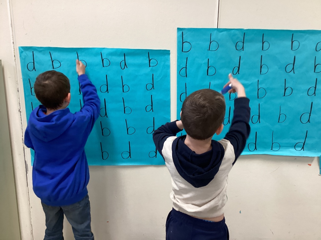 Two students stand at a wall pointing to letters on large blue posters, practicing letter recognition.