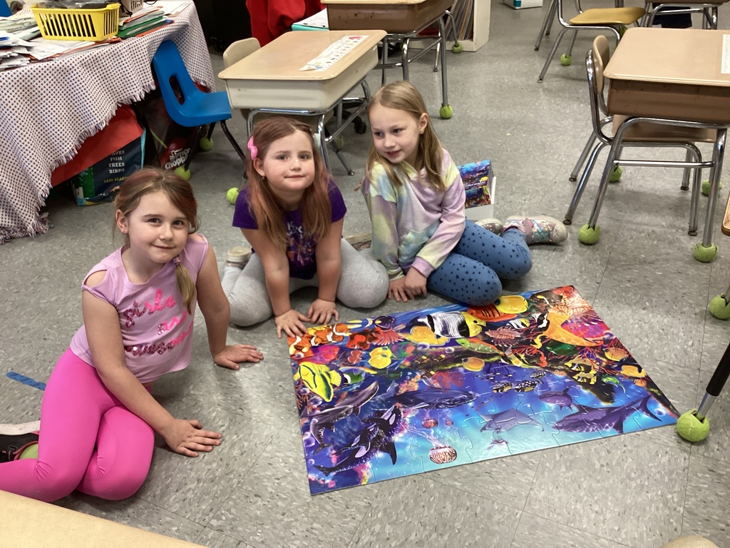 Three young students kneel on the classroom floor, smiling beside a completed ocean-themed jigsaw puzzle.