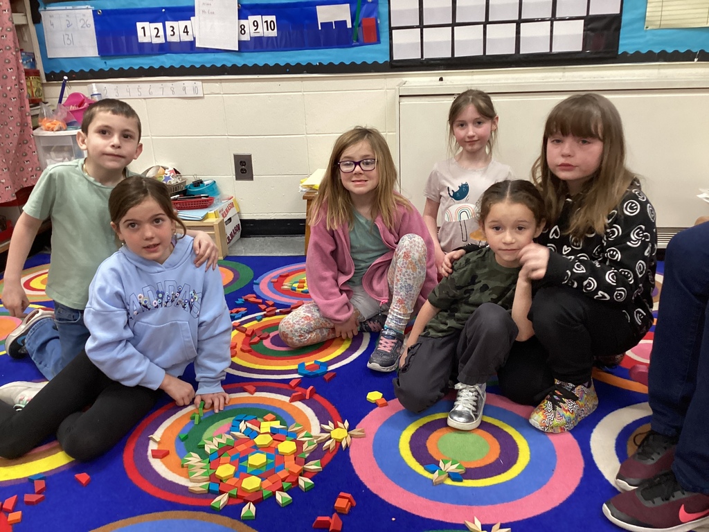 A group of six students sit on a bright rug surrounded by pattern blocks, posing with their collaborative design.