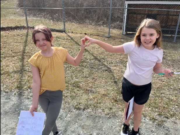 Two girls stand outside holding hands in a heart shape, smiling while one holds a worksheet and the other a notebook.