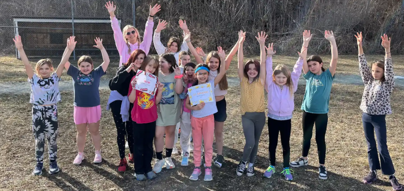A group of elementary-aged girls stands outdoors with arms raised, smiling and celebrating while holding certificates for achievement.