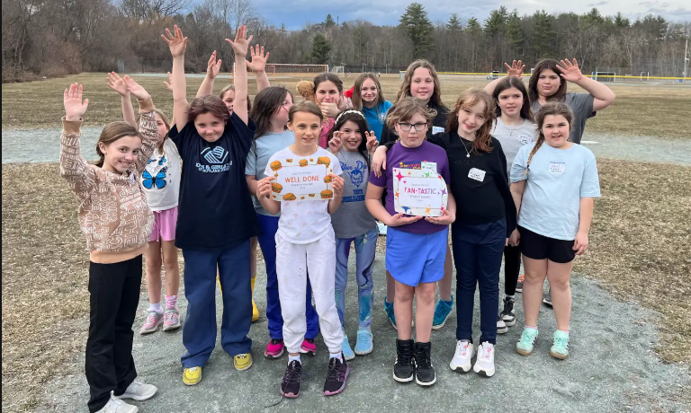 A larger group of girls stands outside posing with raised hands while two students in front hold “Well Done” and “Fan-tastic” award certificates.