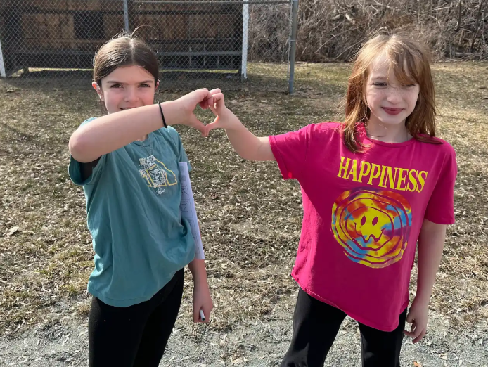 Two girls stand together forming a heart shape with their hands, smiling in a grassy outdoor area.