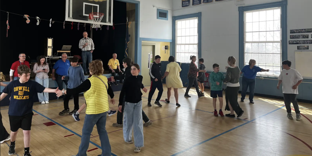 Students and adults rotate partners and step through a coordinated group activity in a gym, with a stage and banners visible behind them.