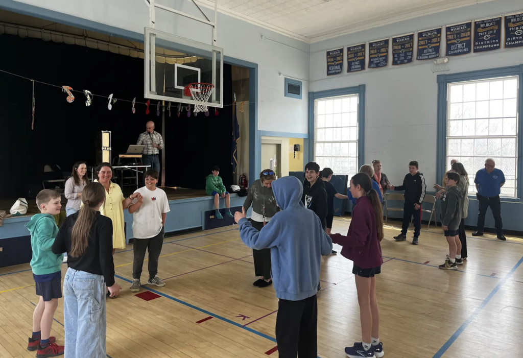 Students and adults stand in a circle in a school gym, holding hands and smiling during a group activity while a presenter stands on a stage behind them.