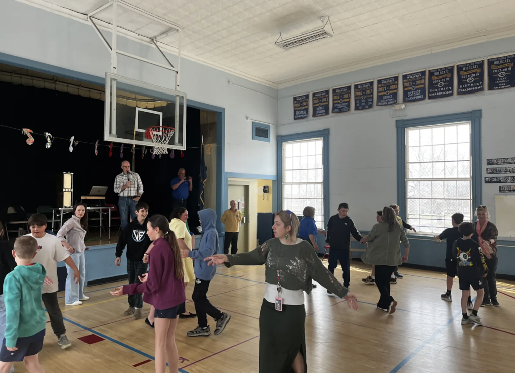 A teacher gestures while guiding students moving in pairs around the gym, with others practicing the same activity in the background.