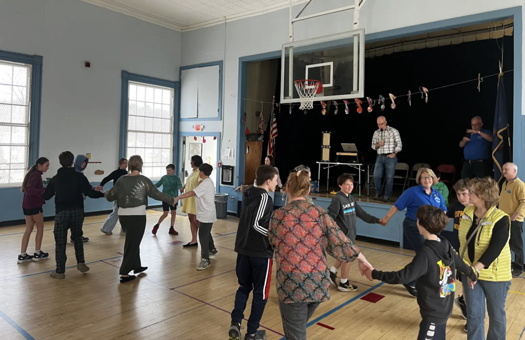 Participants move in a large circle holding hands in a gym, working together in a coordinated group activity while staff observe and assist.