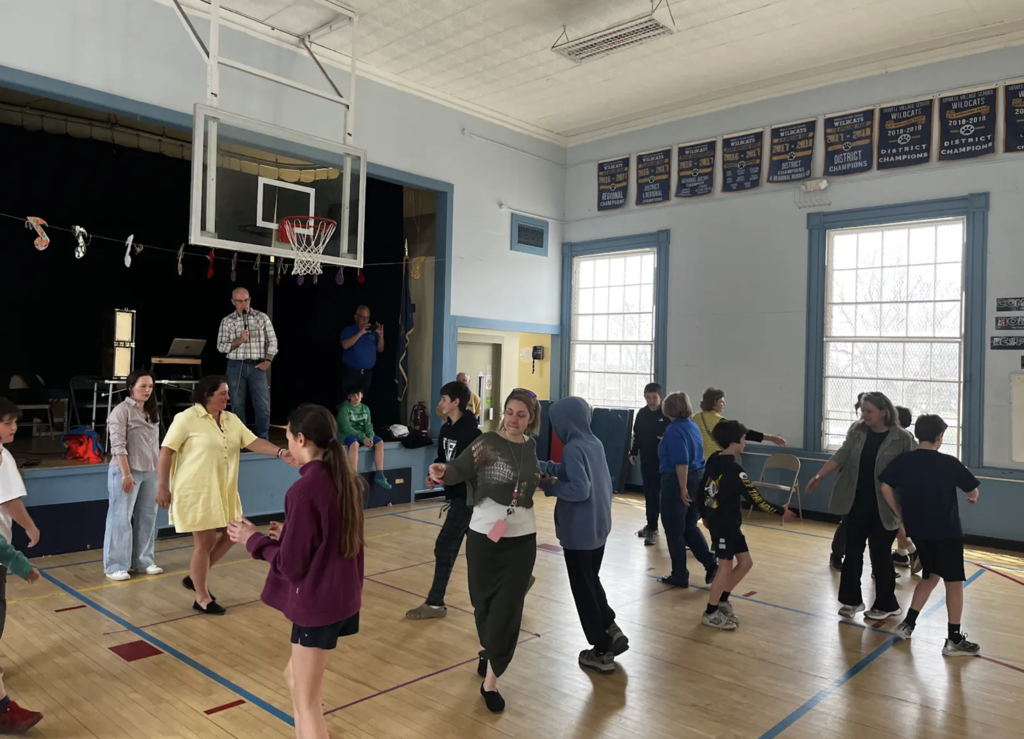 Students and adults walk and turn in pairs across the gym floor during an interactive movement activity, with sunlight coming through large windows.