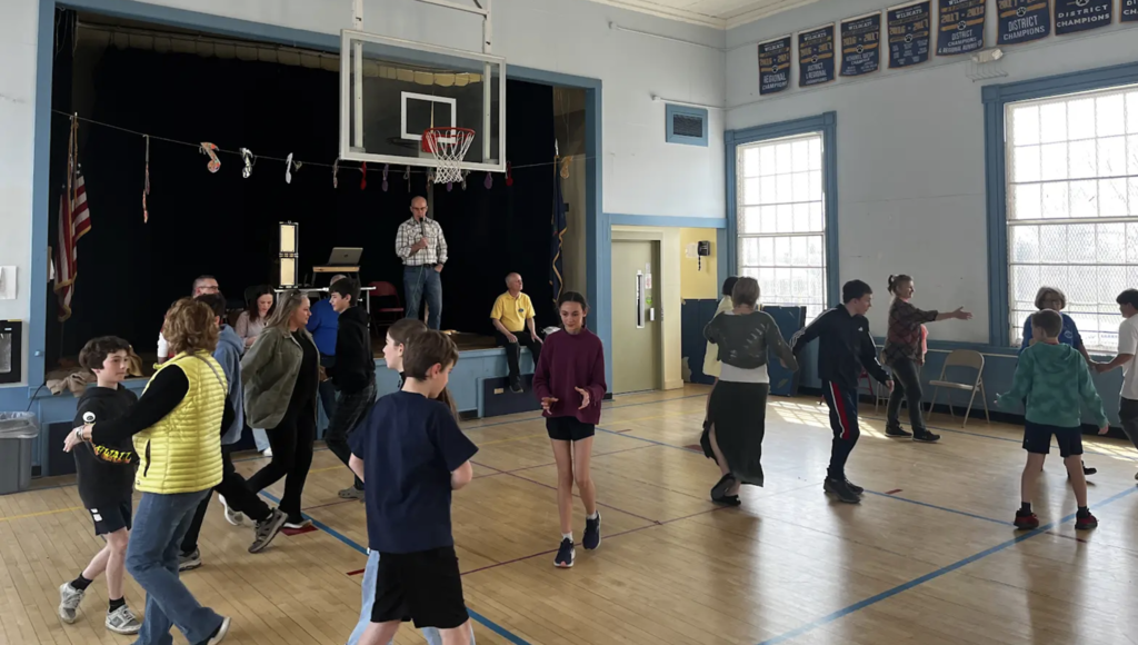 Participants continue a partner-based movement activity in the gym, walking and turning while maintaining spacing across the floor.