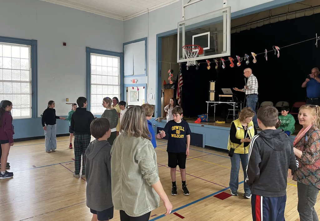 Students and adults pair up across a gym floor, practicing a partner activity as a man speaks from a stage with audio equipment.