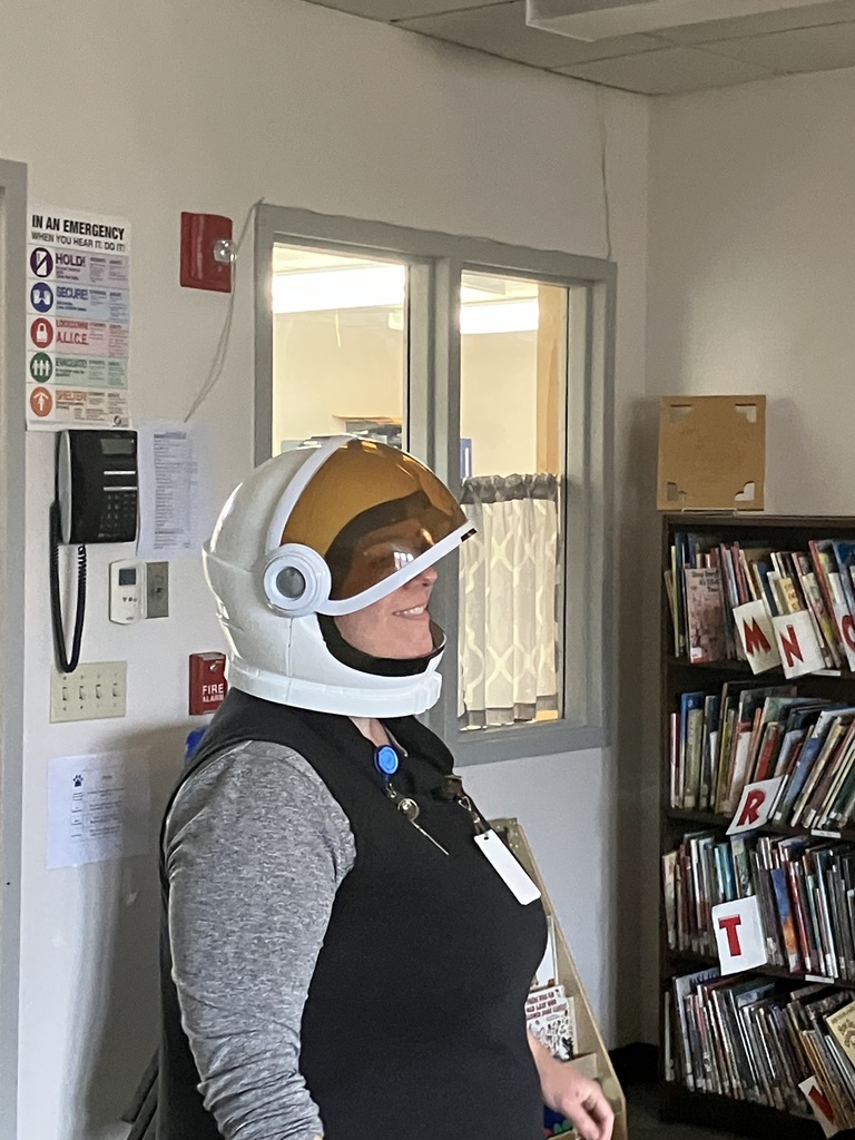  A side profile of an adult smiling while wearing a white astronaut helmet with a gold-tinted visor. They are standing in a classroom or library setting next to a tall bookshelf filled with children's books.