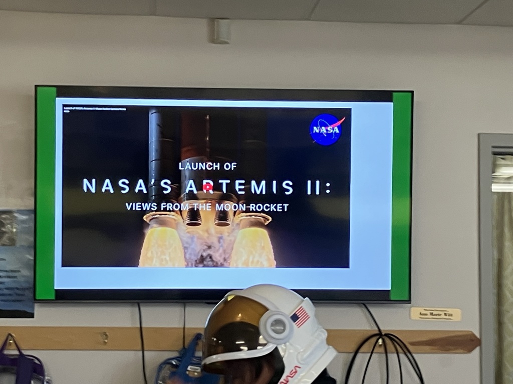  A close-up of a television screen displaying the title "LAUNCH OF NASA'S ARTEMIS II: VIEWS FROM THE MOON ROCKET." Below the screen, the top of a person’s head is visible wearing a white and gold astronaut helmet with a NASA logo.  
