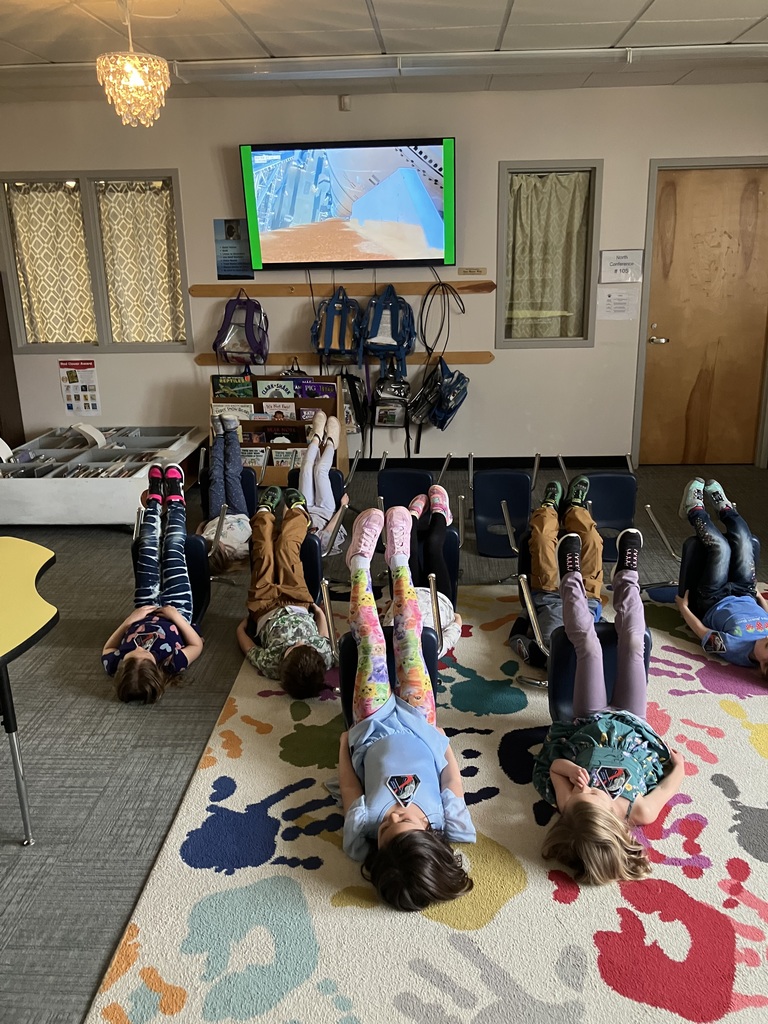  A wide shot of a classroom where several children are lying on their backs with their legs propped up against blue chairs, facing a television screen. The TV shows a simulation of a rocket launch. The room has a colorful rug, a bookshelf, and a chandelier. 