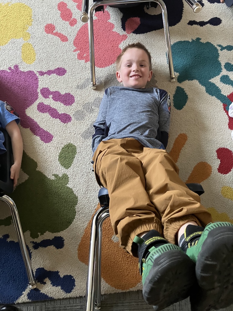  A young boy with a joyful smile lies on a handprint-patterned rug. He is leaning back against a tipped school chair, wearing brown pants and green sneakers, looking directly at the camera. 