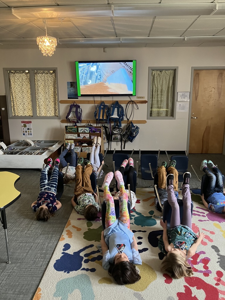  A similar wide shot to the previous image, showing the group of children in their "launch positions" on the floor. The classroom is dimly lit by a crystal chandelier, and backpacks hang on the wall beneath the TV screen.  