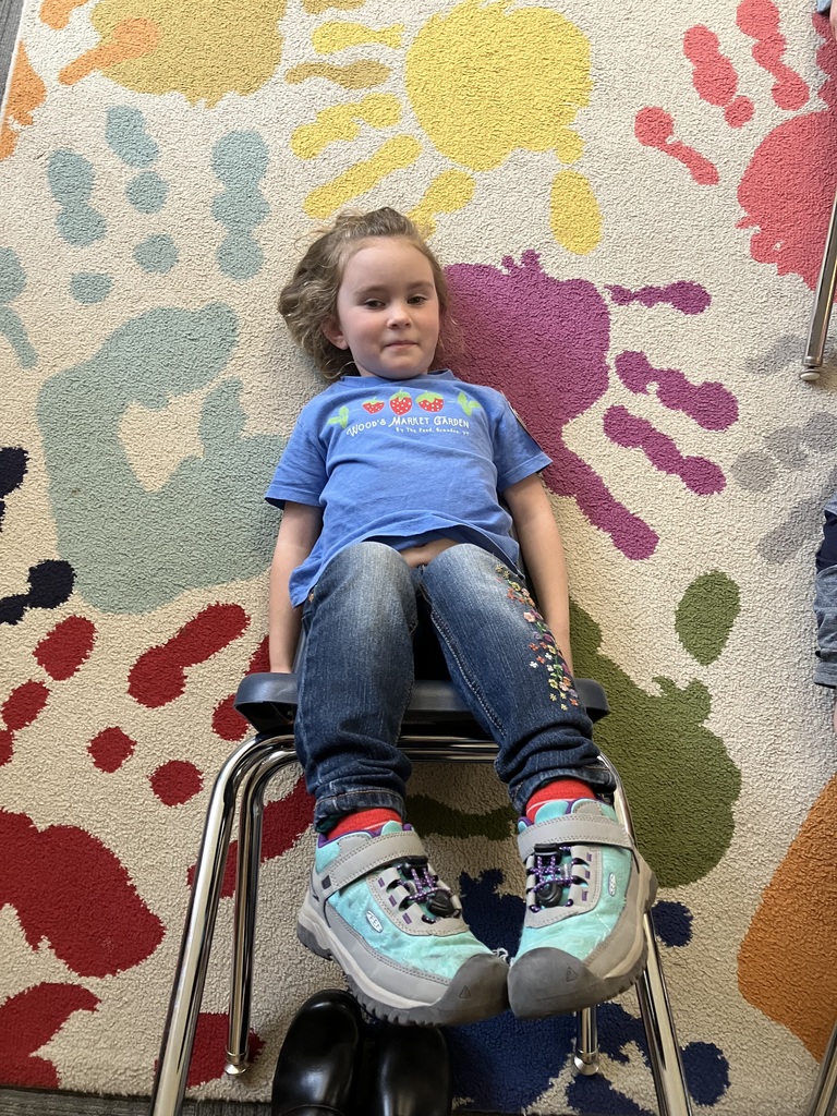 A young child with light brown hair lies on a colorful rug featuring large, bright handprints. The child is resting their back against the seat of a blue chair that has been tipped backward, with their legs extending toward the camera.