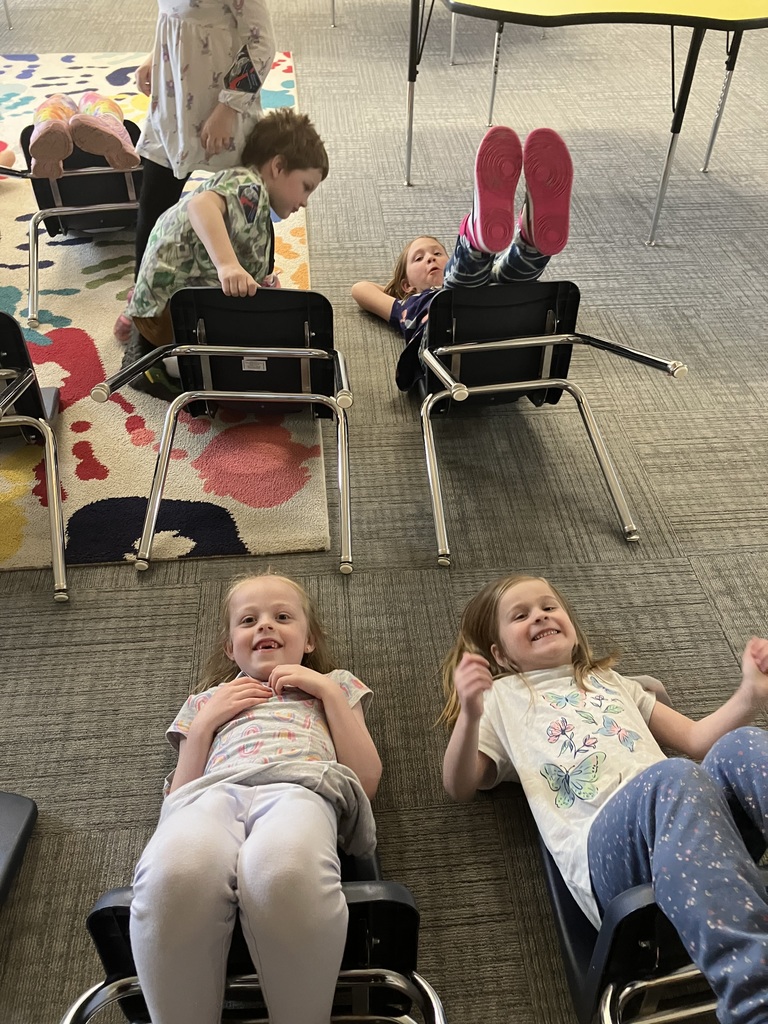 Two young girls lie on their backs on a grey carpeted floor, smiling at the camera. In the background, other children are positioned upside-down on blue school chairs, with their legs in the air, mimicking a rocket launch position.  IMG_3214.jpg A young child with light brown hair lies on a colorful rug featuring large, bright handprints. The child is resting their back against the seat of a blue chair that has been tipped backward, with their legs extending toward the camera.  